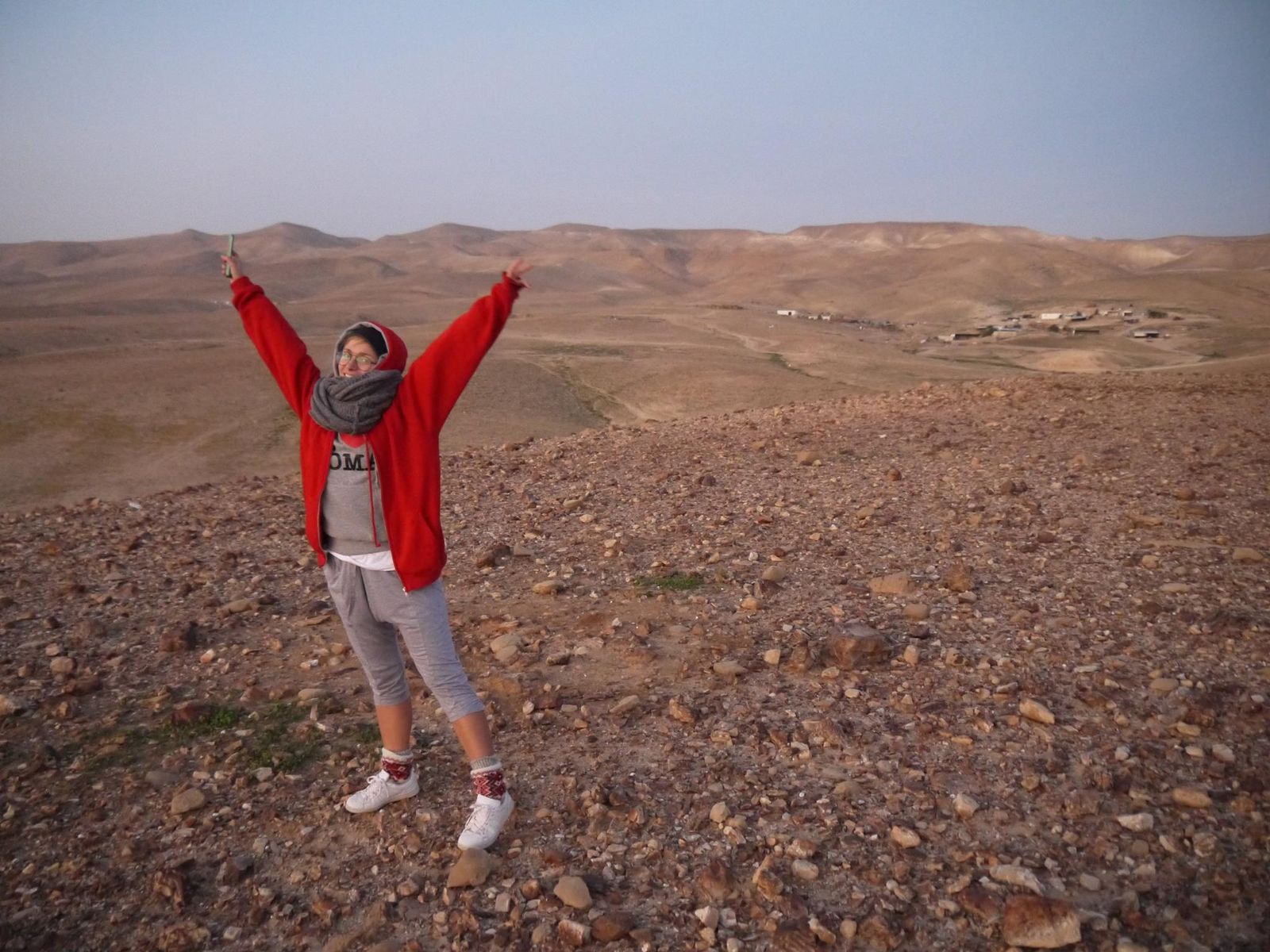   Izrael. Młoda kobieta, ubrana w czerwoną kurtkę, stoi na pustyni. Jest radosna, ma uniesione w górę ręce.   