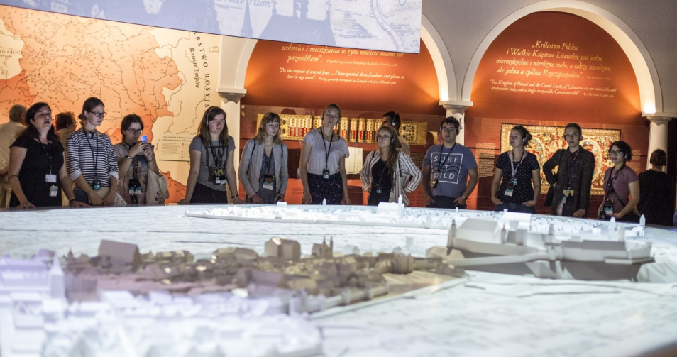 Grupa młodzieży stoi przed ogromną, podświetloną makietą średniowiecznego Krakowa. W tle ściany wystawy "1000 lat historii Żydów polskich" Muzeum POLIN