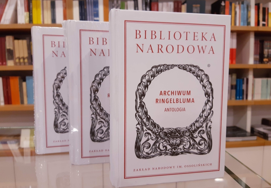 Okładka książki "Archiwum Ringelbuma. Antologia" w wydaniu Biblioteki Narodowej seria "Ossolineu". Ustawiona pionowo na stole. W tle regał z innymi książkami