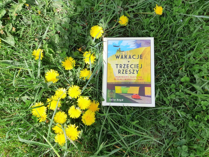 Okładka książki:  Julia Boyd "Wakacje w Trzeciej Rzeszy". Książka leży w trawie, koło kępki kwitnących mleczy