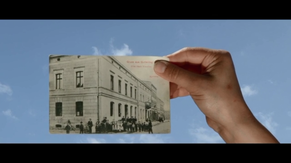 Kadr z filmu "Obłoki płyną nad nami" / "Under a Placid Sky" – film Anny Konik. Widać dłoń, która trzyma czarno-białą fotografię budynku i ulicy. W tle pogodne niebo z obłokami.
