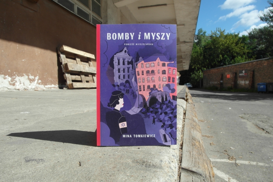 Na betonowej powierzchni stoi książka "Bomby i myszy". W tle frgament starego budynku.