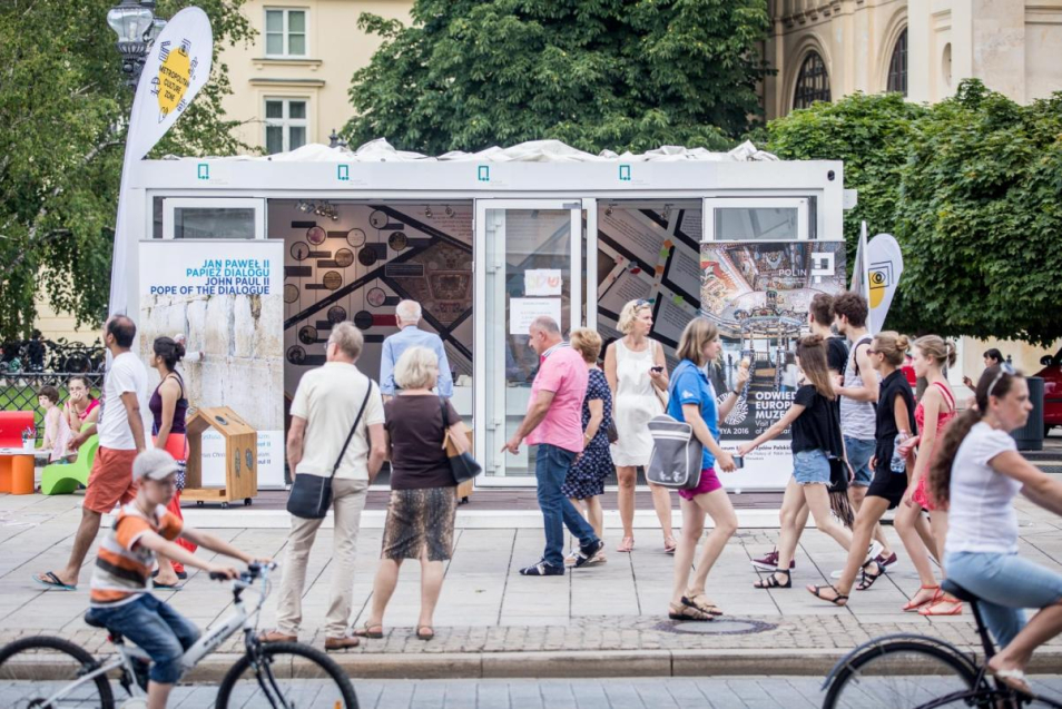 Pawilon "Muzeum na kółkach" stoi w parku miejskim. Wokół gromadzą się ludzie. Część po prostu przechodzi obok.