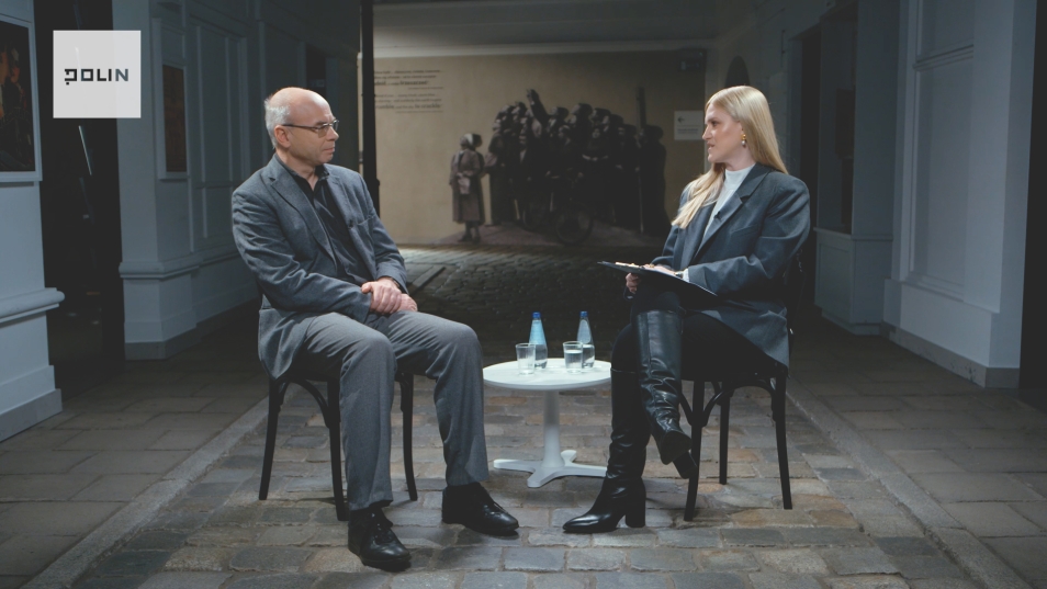 Na zdjęciu, w ciemnym wnętrzu (wystawa Muzeum POLIN) naprzeciw siebie na krzesłach siedzą dwie osoby: mężczyzna w garniturze (prof. Dariusz Stola) i kobieta (Hanna Rydlewska)