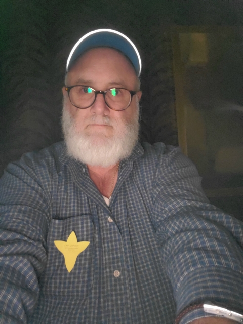 In Polish: mężczyzna z długą siwą brodą, w czapce z daszkiem, robi selfie. Do szarek koszuli ma przypięty papierowy żonkil, symbol akcji Żonkile