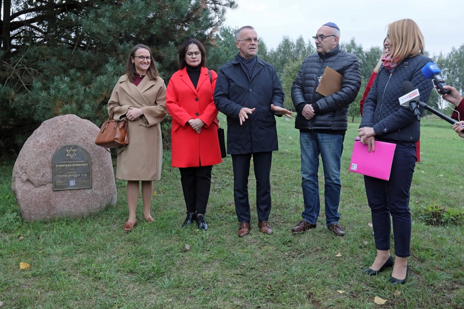 Obok dużego kamienia z plakietką, oznaczającą żydowski cmentarz w Orli, stoi grupa osób. W środku wiceminister Jarosław Sellin oraz dyrektor Muzeum POLIN Zygmunt Stępiński