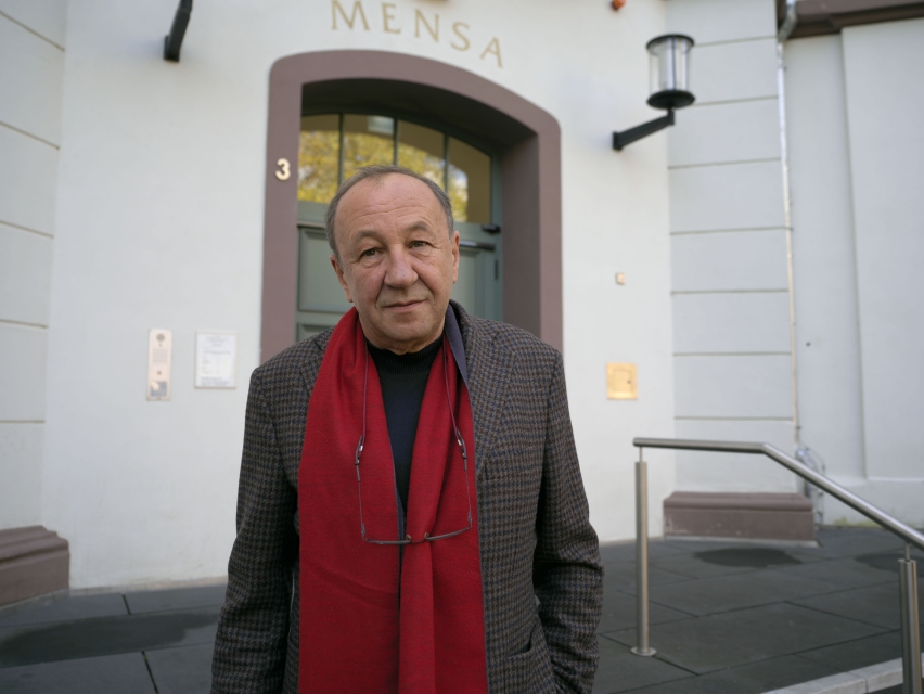 Robert Traba. Mężczyzna w płaszczu i w czerwonej marynarce. Stoi przed budynkiem z napisem Mensa.