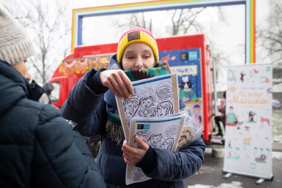 Kobieta w żółtej czapce rozdaje kolorowanki. Nieopodal stoi czerwony chanukobus - przy Zachęcie.