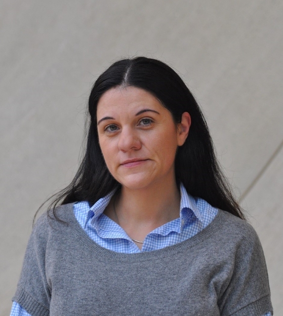 Katharina Friedla. Długowłosa brunetka w niebieskiej koszuli z kołnierzykiem i szarym sweterku.