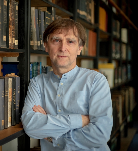 Marcin Wodziński. Brunet w jasnoniebieskiej koszuli. W tle biblioteczka pełna książek.