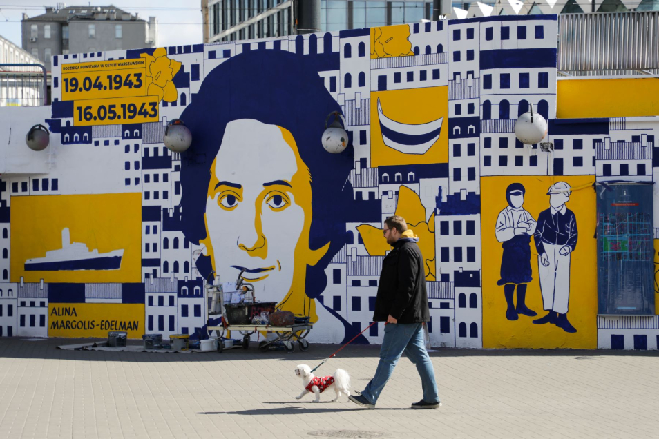 Mężczyzna spaceruje z psem po tzw. warszawskiej patelni, na ścianie mural upamiętniający bohaterki i bohaterów powstania w getcie warszawskim.