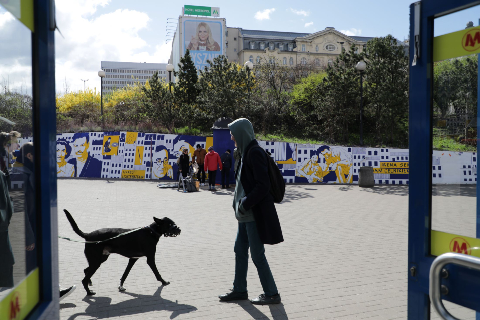 Mężczyzna stoi z psem przy wejściu na stację metra Centrum. W tle, na ścianie okalającej plac, mural upamiętniający bohaterki i bohaterów powstania w getcie warszawskim.