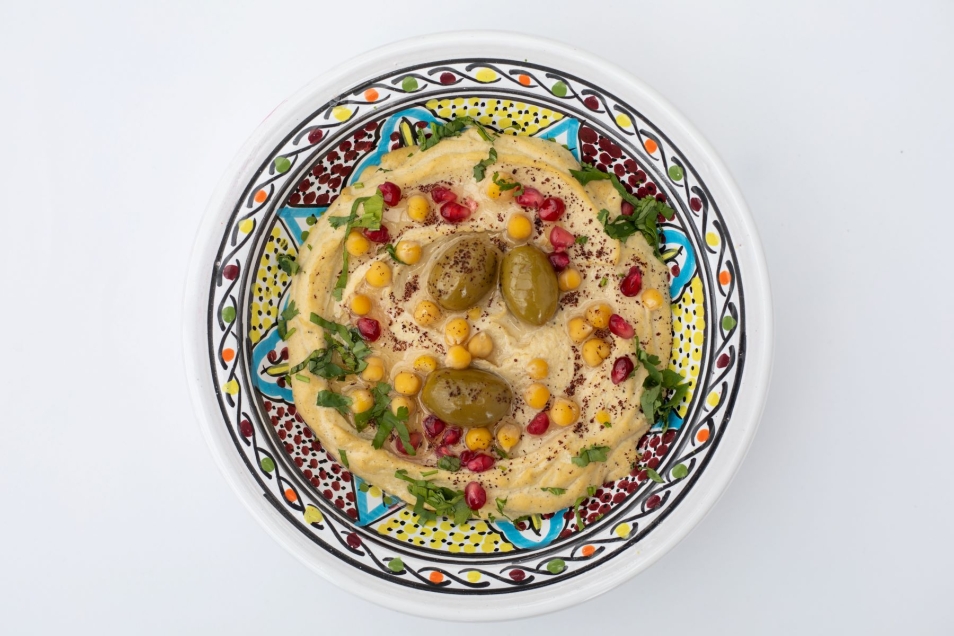 Hummus w kolorowej miseczce. Jest udekorowany oliwkami, ziołami i ciecierzycą.