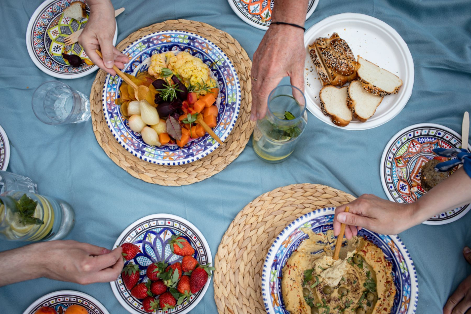 Kilka potraw stoi na kocu. Są tam truskawki, hummus, chałka. Sięga po nie kilka osób.