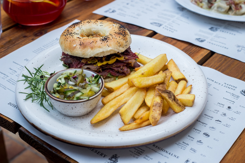 Na stole w restauracji stoi talerz z kanapką a bajgla. Obok bajgla pojemniczek z sałatką i frytki.