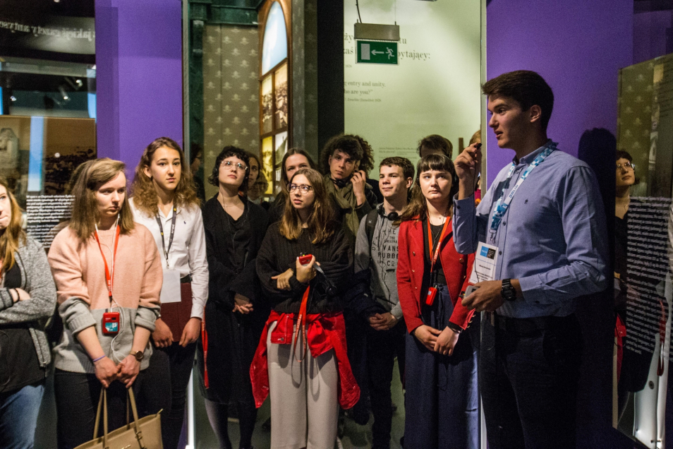 Młody chłopak oprowadza grupę rówieśników po wystawie stałej w Muzeum POLIN. Mówi przez mikrofon.