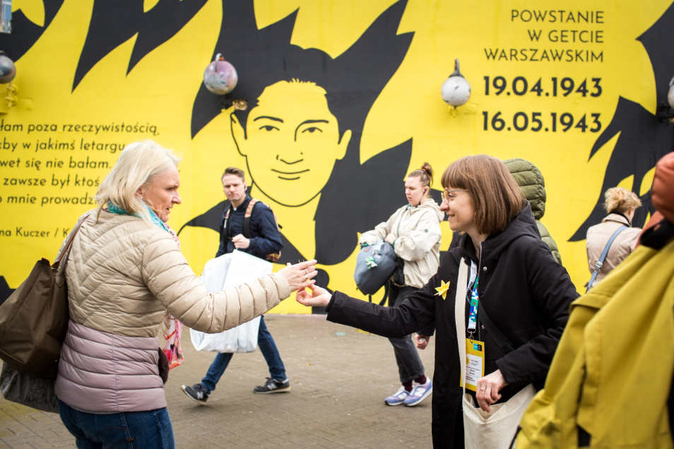 Wolontariuszka wręcza papierowy żonkil kobiecie przy metrze Centrum. W tle mural upamiętniający powstanie w getcie warszawskim.