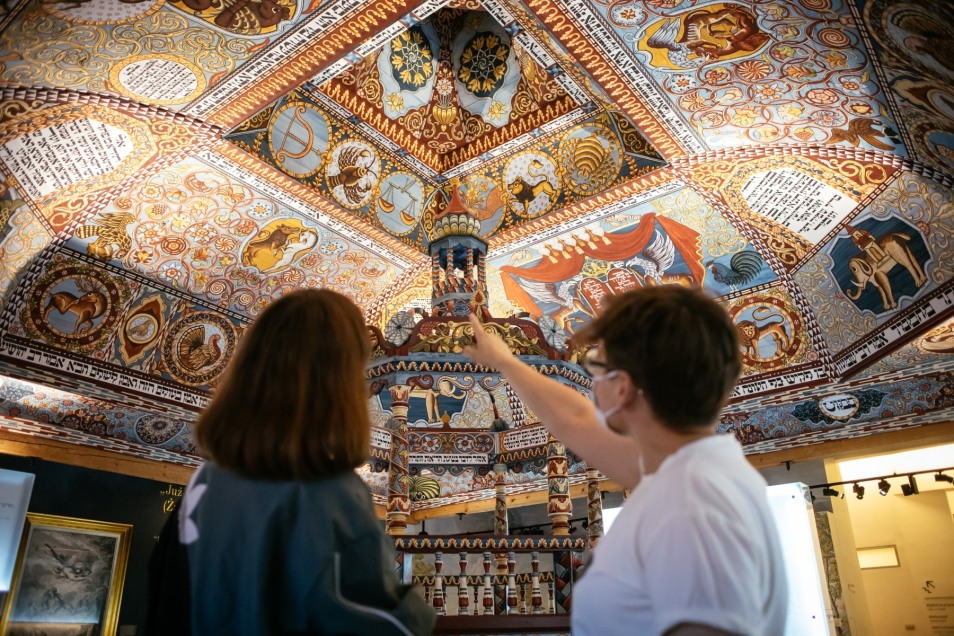 Dwoje zwiedzających - dziewczyna i chłopak - stoi przy rekonstrukcji synagogi w Muzeum POLIN. Chłopak wskazuje palcem kopułę drewnianej bimy.