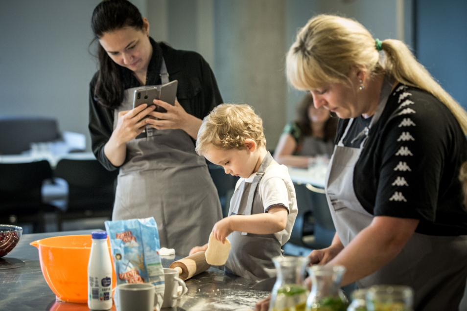 A boy and a woman are taking part in culinary workshops. Some woman is standing next to them and taking a photo of a boy.