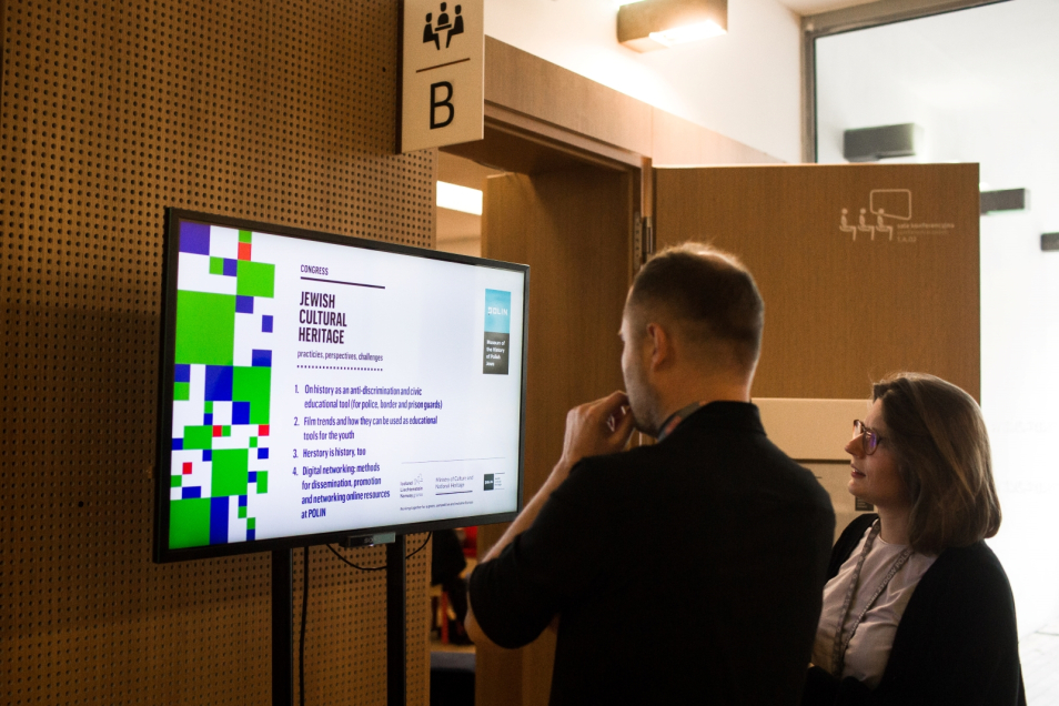 Dwoje uczestników kongresu stoi przed salą konferencyjną i patrzy na ekran multimedialny z programem wydarzenia.