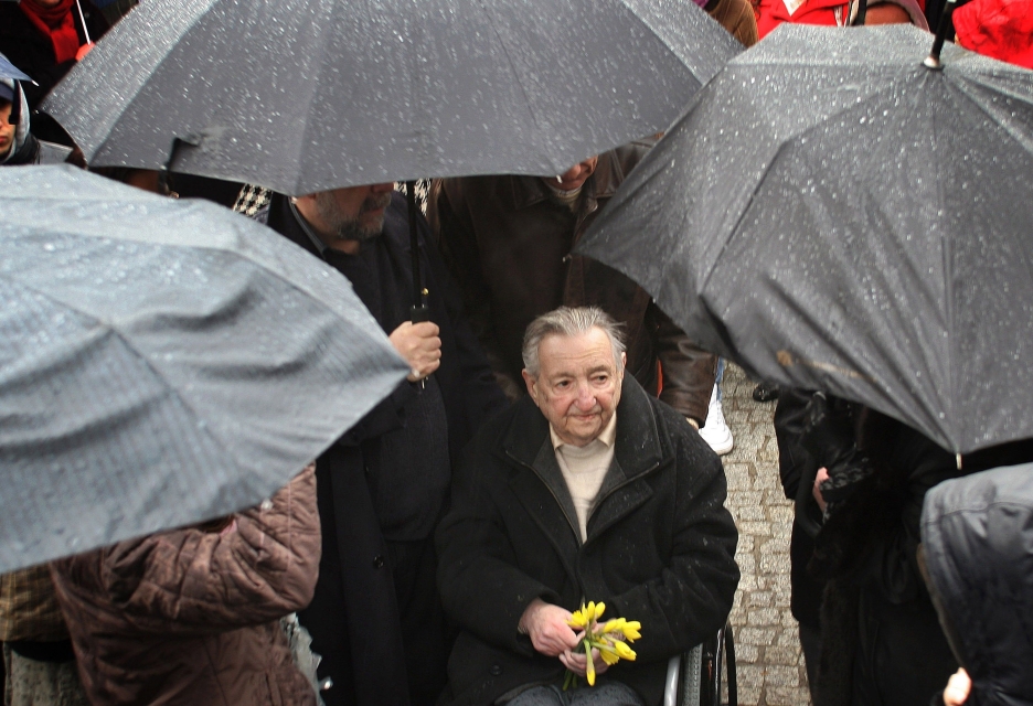 Marek Edelman stoi z odkrytą głową w deszczu. W ręku trzyma kilka żonkili. Wokół niego osoby ukryte pod parasolami. 65. rocznica powstania w getcie warszawskim, przy Kopcu Anielewicza
