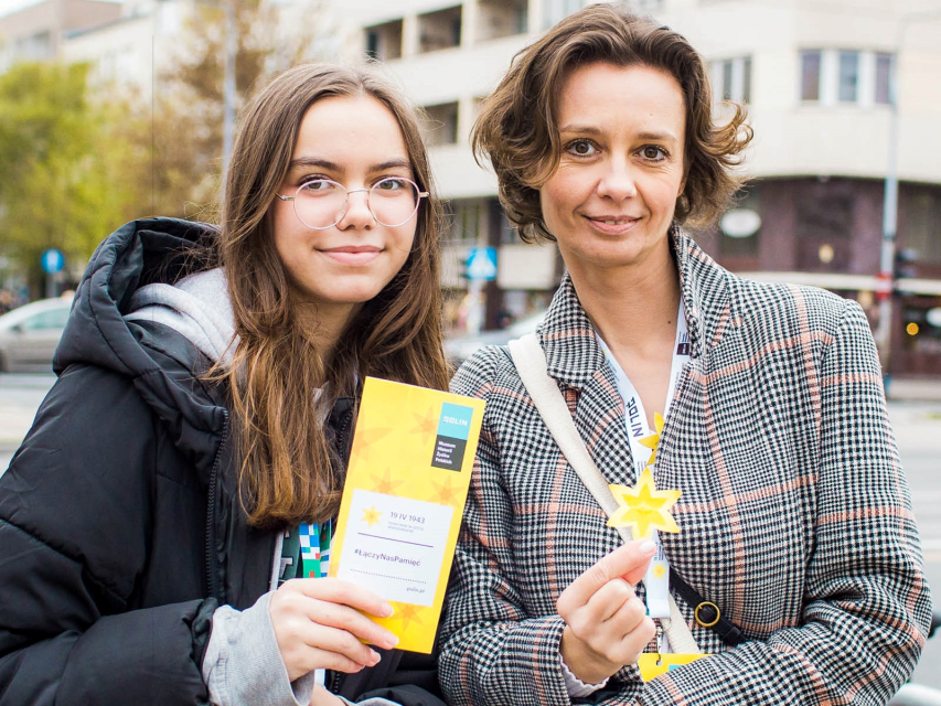 Dwie uśmiechnięte wolontariuszki - nastoletnia i dorosła. Jedna trzyma ulotkę o akcji Żonkile, a druga papierowego żonkila.