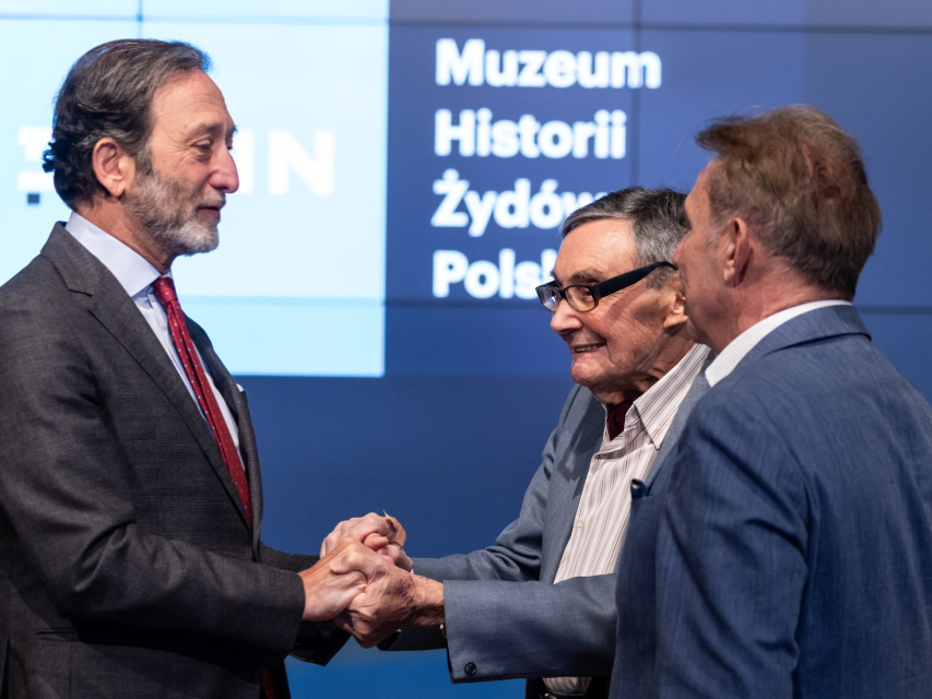 Marian Turski i ambasador Niemiec Viktor Elbling ściskają się za dłonie. Obok nich stoi Piotr Wiślicki.