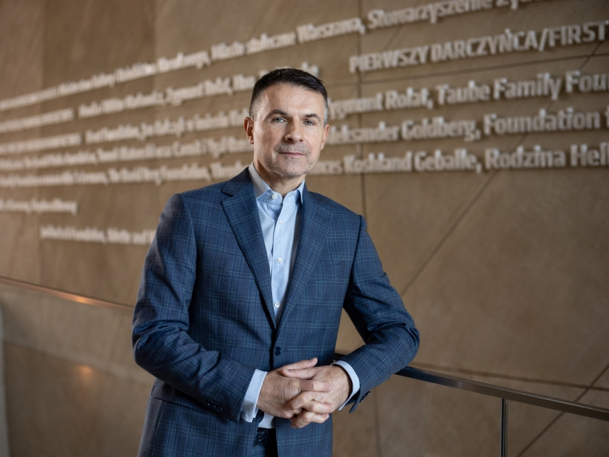 Robert Wereda opiera się o poręcz mostku w holu głównym Muzeum POLIN. W tle ściana z nazwiskami darczyńców.