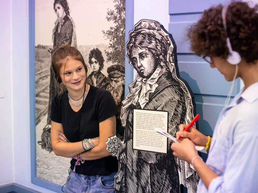 Wystawa stała w Muzeum POLIN. Dziewczyna patrzy na chłopaka, który coś notuje. Oboje są w słuchawkach.