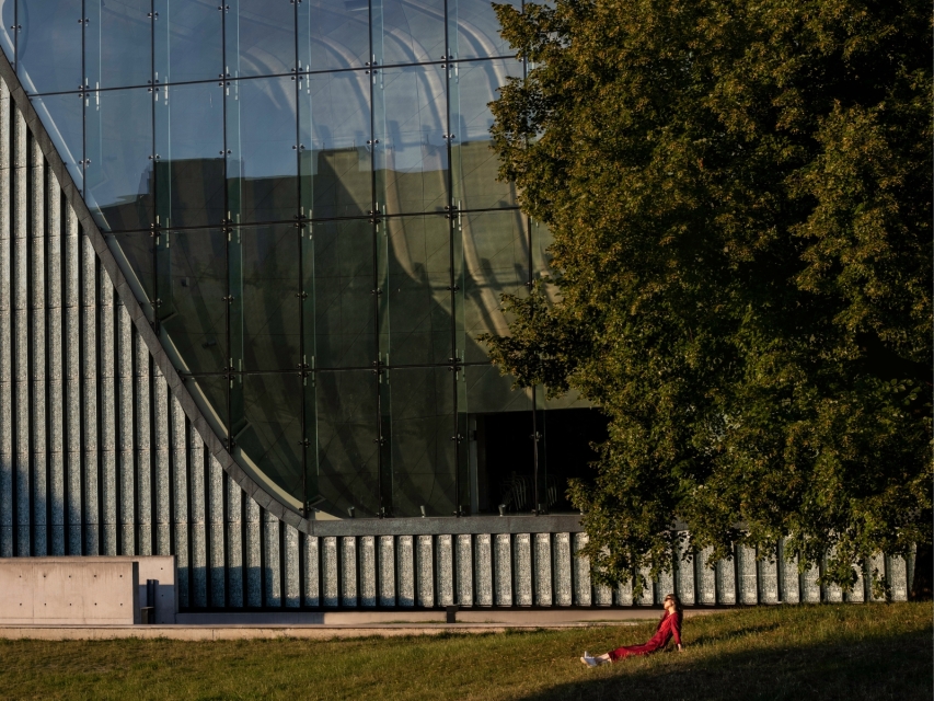 Dziewczyna siedzi pod drzewem przy budynku Muzeum POLIN.