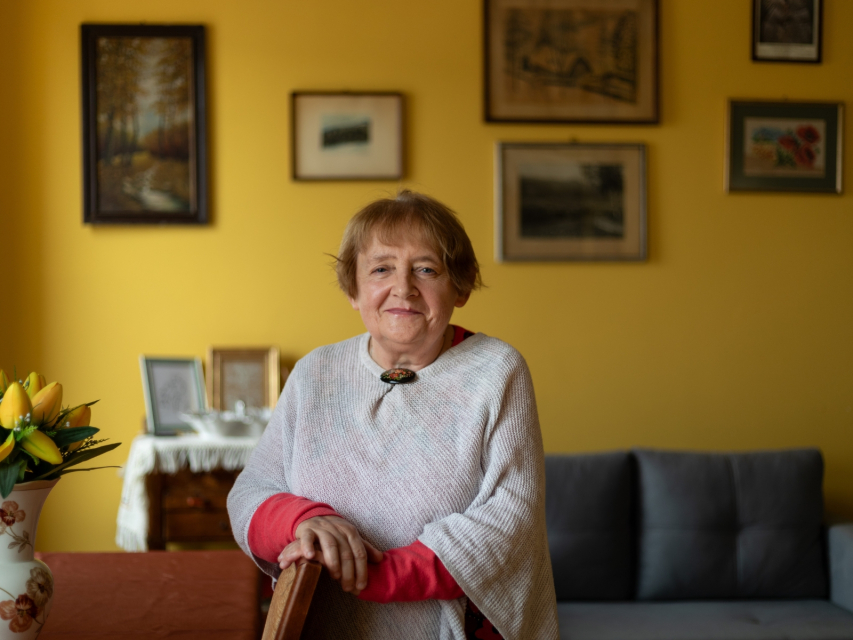 Ewa Wroczyńska w mieszkaniu. Na żółtej ścianie wisi kilka obrazków.