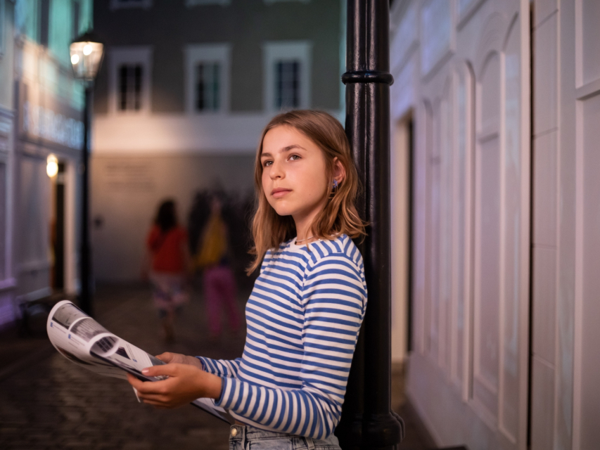 Dziewczyna stoi oparta o lampę na wystawie stałej w POLIN, w galerii "Na żydowskiej ulicy". Trzyma jakieś kartki w dłoniach.