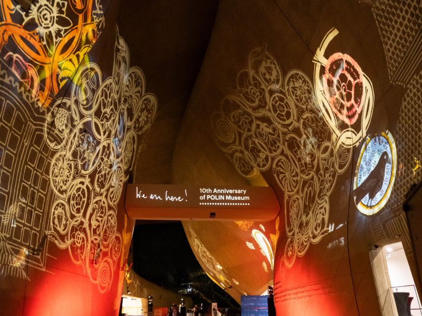 Hol główny Muzeum POLIN podświetlony z okazji 10. urodzin. Na ścianach kolorowe animacje, a na mostku napis We are here! 10th anniversary of POLIN Museum.
