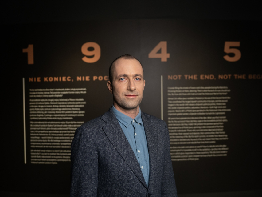 Kamil Kijek na tle planszy z tytułem wystawy "1945. Nie koniec, nie początek".