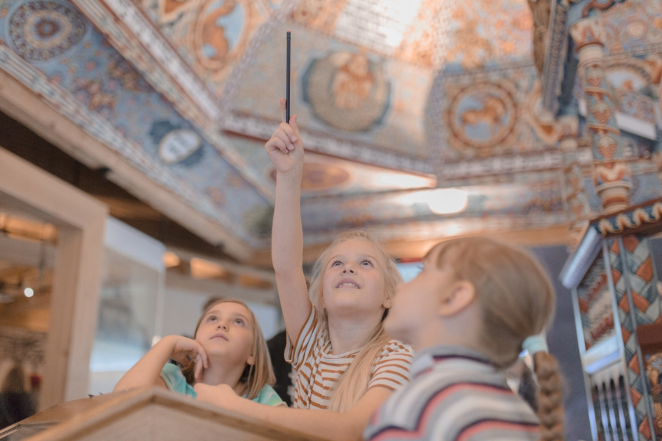 Trzy dziewczynki opierają się o stół na wystawie stałej w Muzeum POLIN, na którym stoi figurka gęsi, a z góry zwisa sztuczny czosnek.