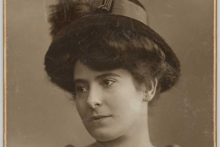 Fotografia portretowa Heleny Rubinstein w półprofilu. Kobieta jest ubrana w bluzkę z delikatnym wzorem, z dekoltem w kształcie litery V, wykończonym niewielkim bukietem żywych rumianków. Głowę przystraja kapelusz z wąskim, zaokrąglonym w dół rondem, z szeroką wstążką.