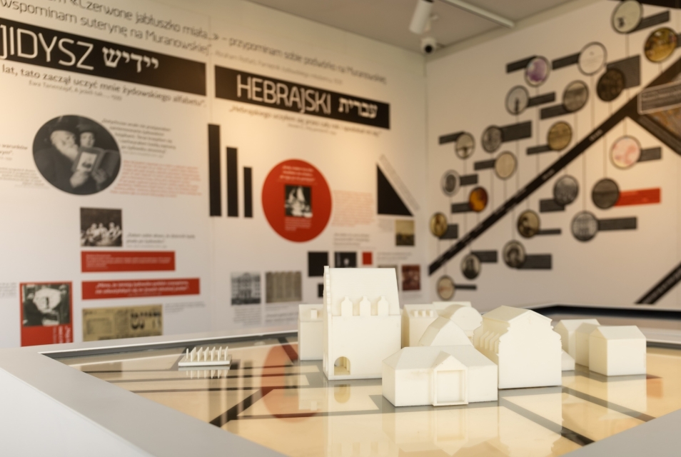 Fragment wystawy "Muzeum na kółkach". Domki ustawione na makiecie. W tle plansze z napisami jidysz i hebrajski.