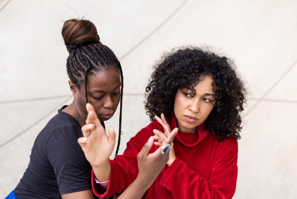 Two dark-skinned women are standing next to each other. They are holding their hands up, close to their faces.