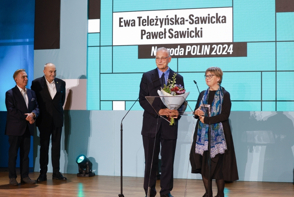 Ewa Teleżyńska-Sawicka and Paweł Sawicki stand on stage during 2024 POLIN Award gala. Next to them, Zygmunt Stępiński and Piotr Wiślicki stand in the corner.