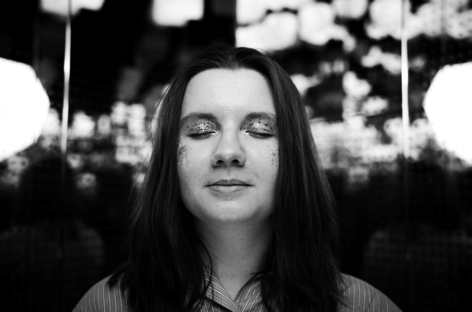 The girl has her eyes closed and shiny eyeshadow on her eyelids. She poses against a backdrop of houses hanging from the ceiling in the Bonds gallery.