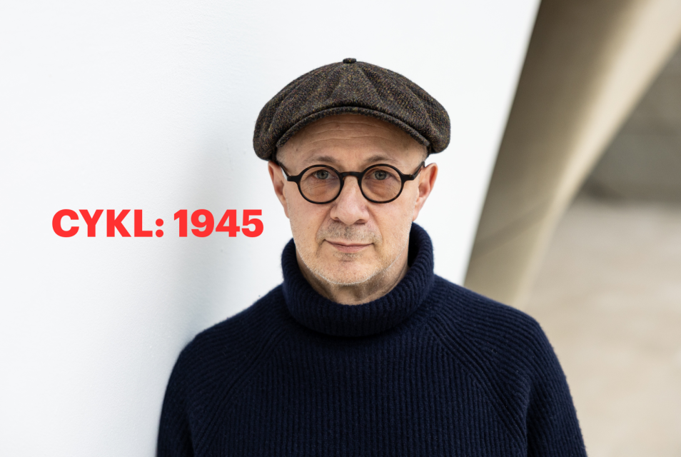 Mikołaj Grynberg stoi w holu głównym Muzeum POLIN. Ma kaszkiet i okulary. Po lewej stronie czerwony napis cykl "1945".