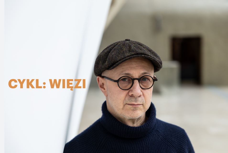 Mikołaj Grynberg stoi w holu głównym Muzeum POLIN. Ma okulary i kaszkiet. Obok złoty napis cykl: "Więzi".