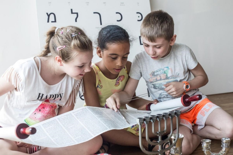 Troje dzieci ogląda zwój Tory na warsztatach edukacyjnych. Za nimi stoi tablica z alfabetem hebrajskim.