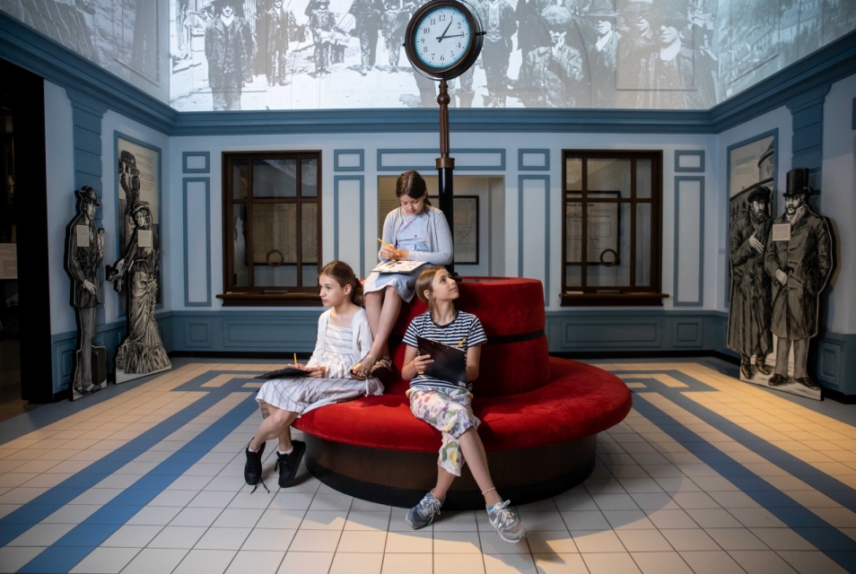 Trzy dziewczynki siedzą na kanapie w galerii "Wyzwania nowoczesności" na wystawie stałej w Muzeum POLIN. W dłoniach trzymają podkładki na kartki i ołówki.