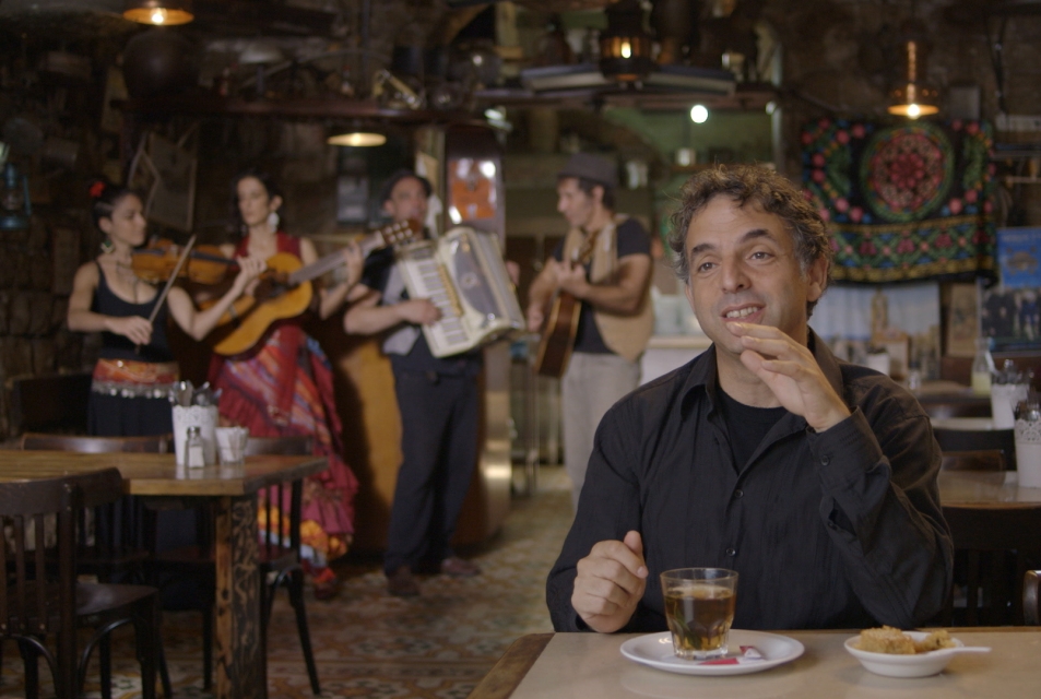 Kadr z filmu "Etgar Keret. Oparte na prawdziwej historii". Keret siedzi przy stole, pije herbatę, w tle zespół z instrumentami.