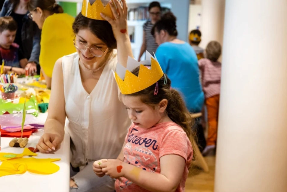 A woman and a girl are sitting at a table full of colourful cut-outs. Both are wearing crowns on their heads.