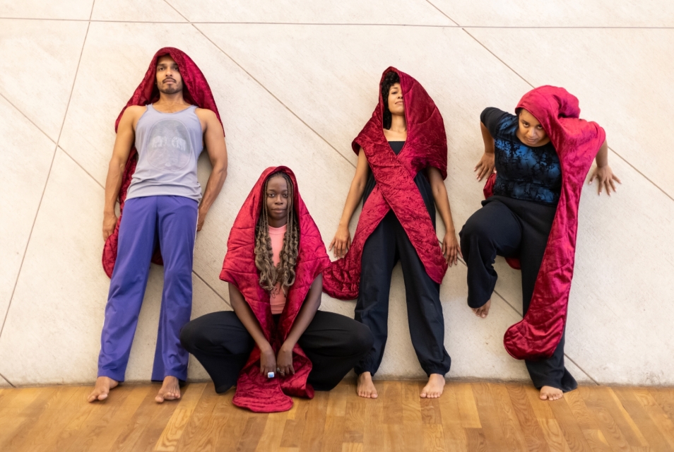 Four dark-skinned people are standing by the wall in the POLIN Museum. They all have red hoods on their heads.