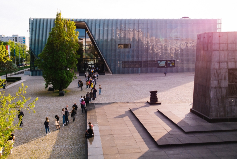 A queue of people is waiting to enter the POLIN Museum building. The sun is shining. The Monument to the Ghetto Heroes is visible on the right.