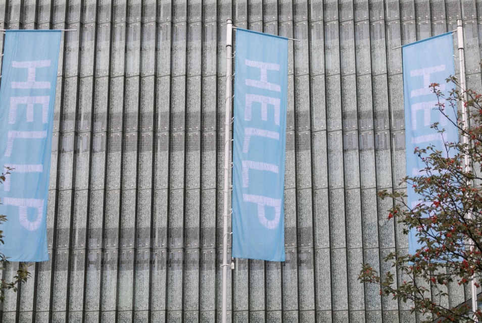 Trzy błękitne flagi z białym napisem HELLP każda. Gest artystyczny Moniki Drożyńskiej. W tle szklane panele ściany Muzeum POLIN (1)