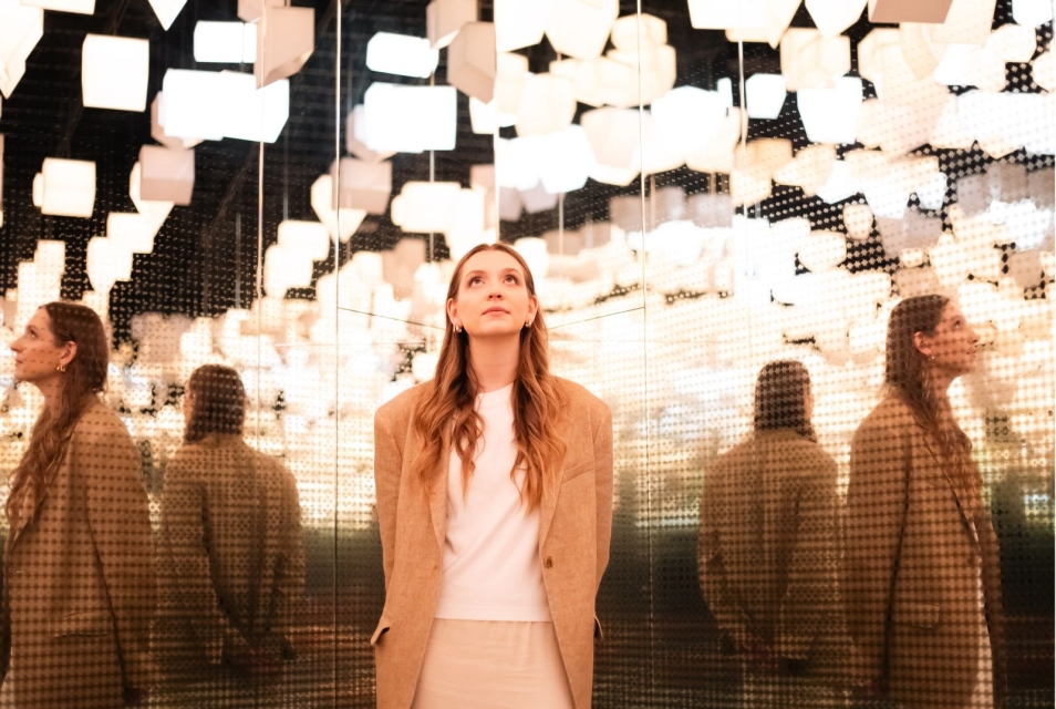 Kobieta z uniesioną głową stoi w galerii "Więzi" pod sufitem wykonanym z wiszących lamp w kształcie domków